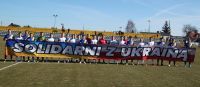Zdjęcie: Piłkarze stojący na boisku sportowym z banerem "Solidarni z Ukrainą"