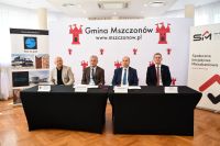 Zdjęcie: mężczyźni podpisujący umowę przy stole, w tle ścianka promująca gminę Mszczonów