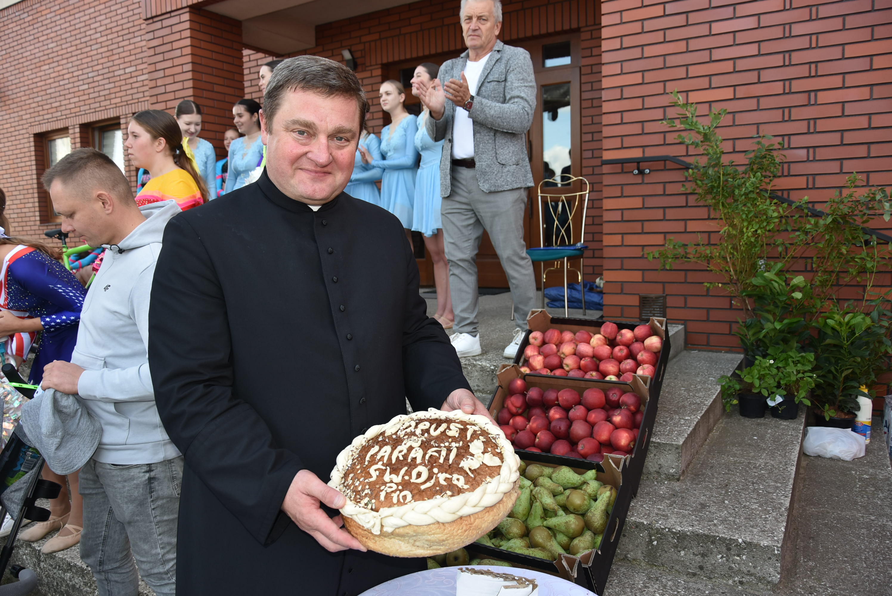 Zdjęcie: ksiądz trzymający koszyk z jabłkami, w tle uczestnicy odpustu parafialnego